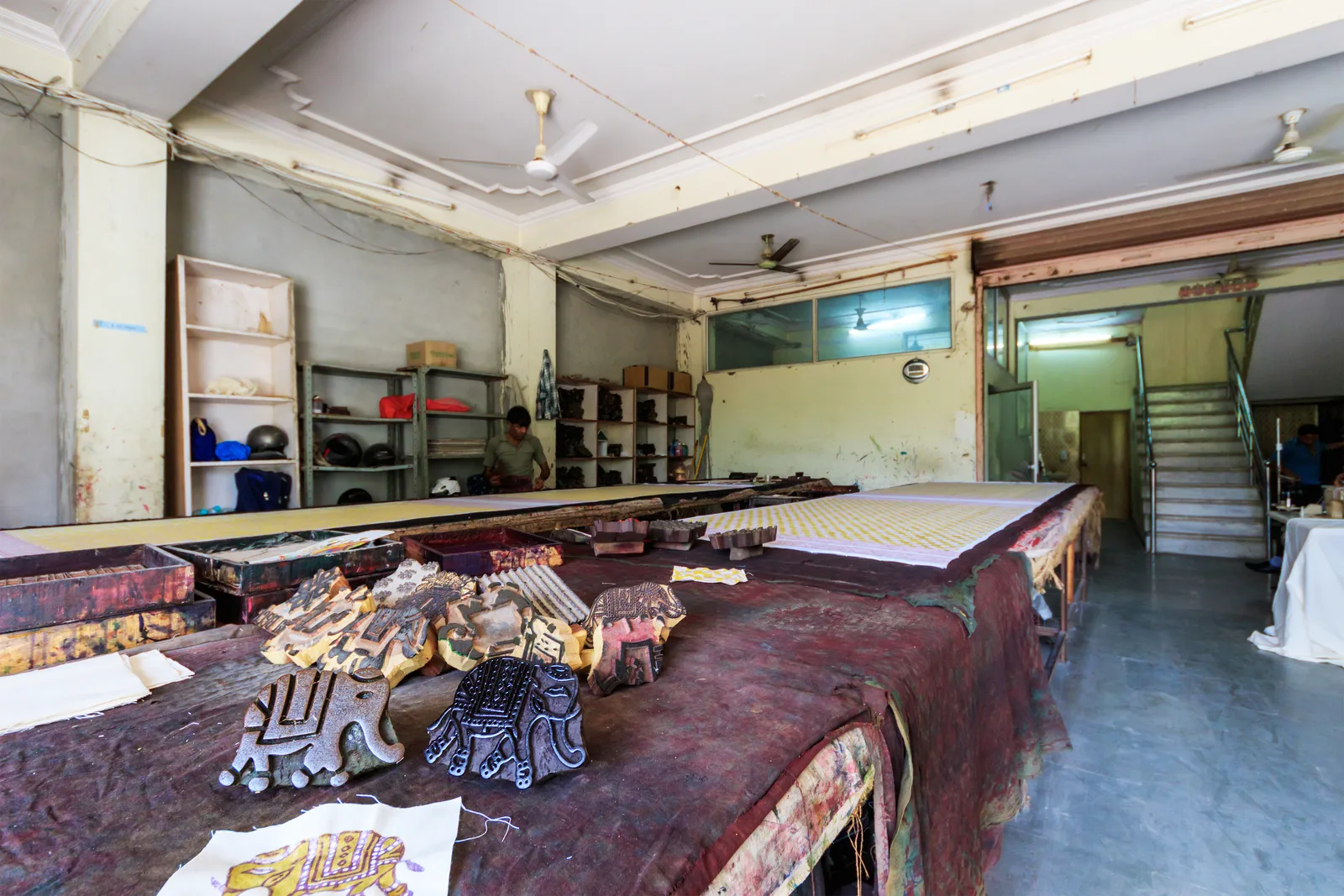 Long padded printing tables inside a Jaipur textile workshop, with stacks of carved wooden blocks and printed cloth laid out