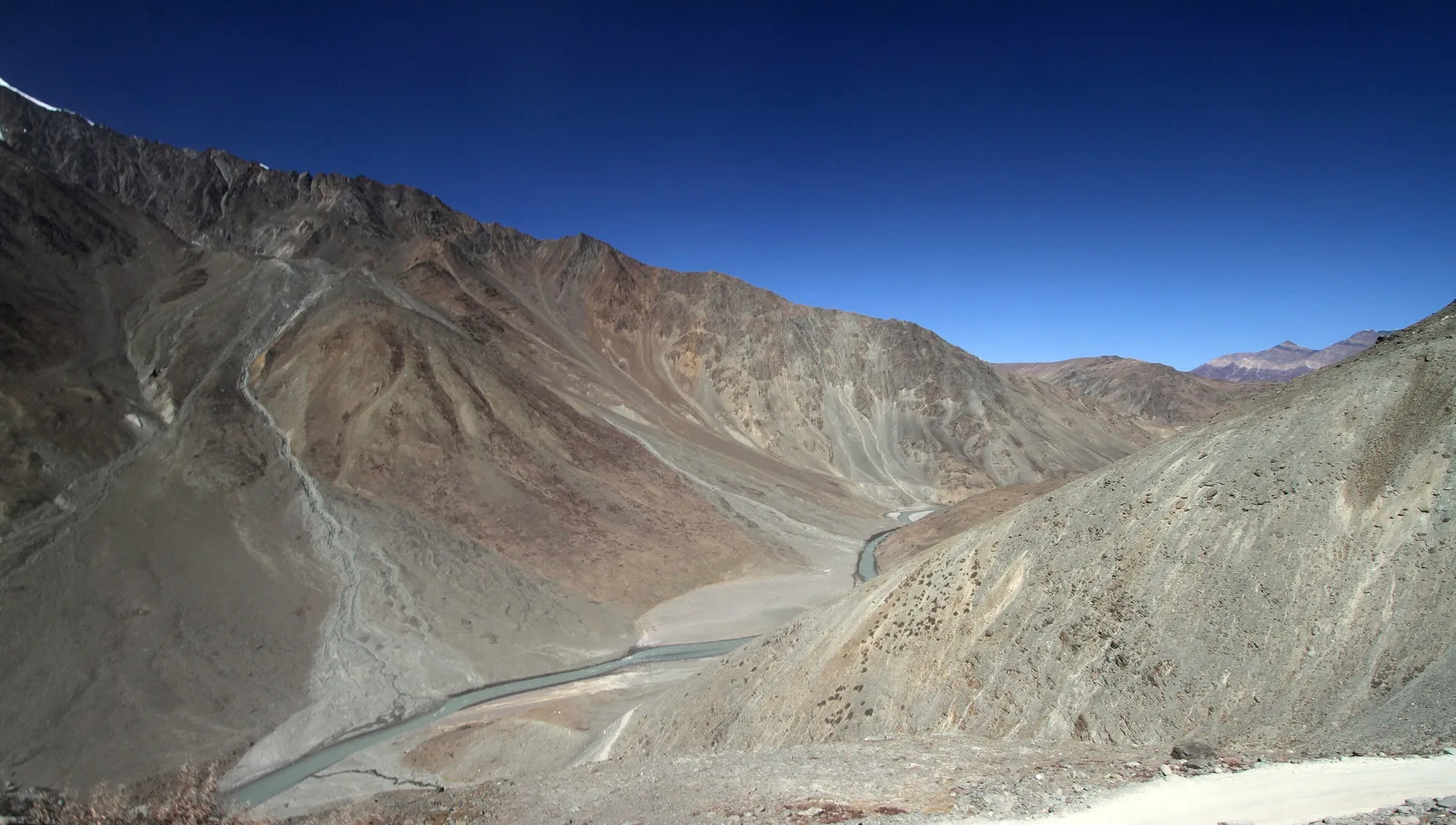 The Chandra river winding through a bare, steep-sided valley below Kunzum Pass on the Lahaul side, with a gravel road in the foreground