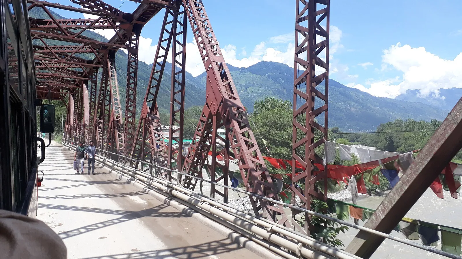 The red steel truss bridge at Bhuntar over the Beas, with prayer flags on the railing and forested hills behind