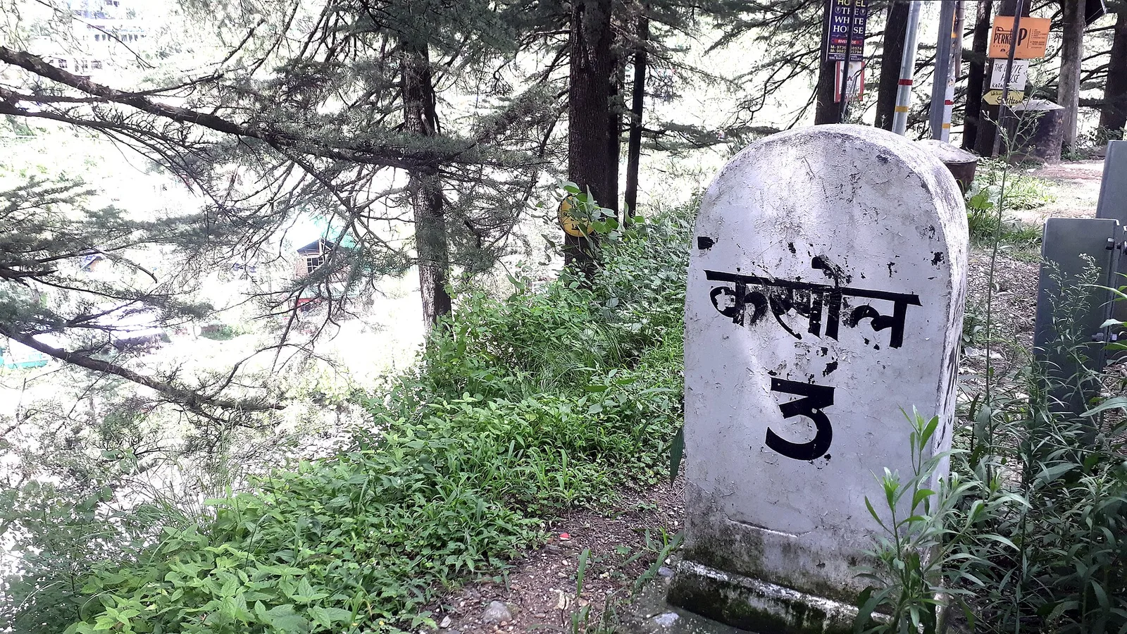 A white roadside milestone reading Kasol 3 in Hindi beside the Parvati valley road, with pine trees and the river valley behind