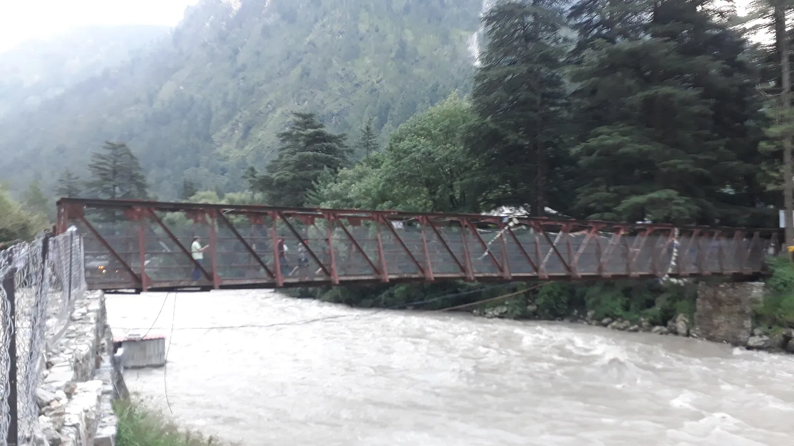 A red steel footbridge over the fast-flowing Parvati river at Kasol, with pine-covered hills and low cloud
