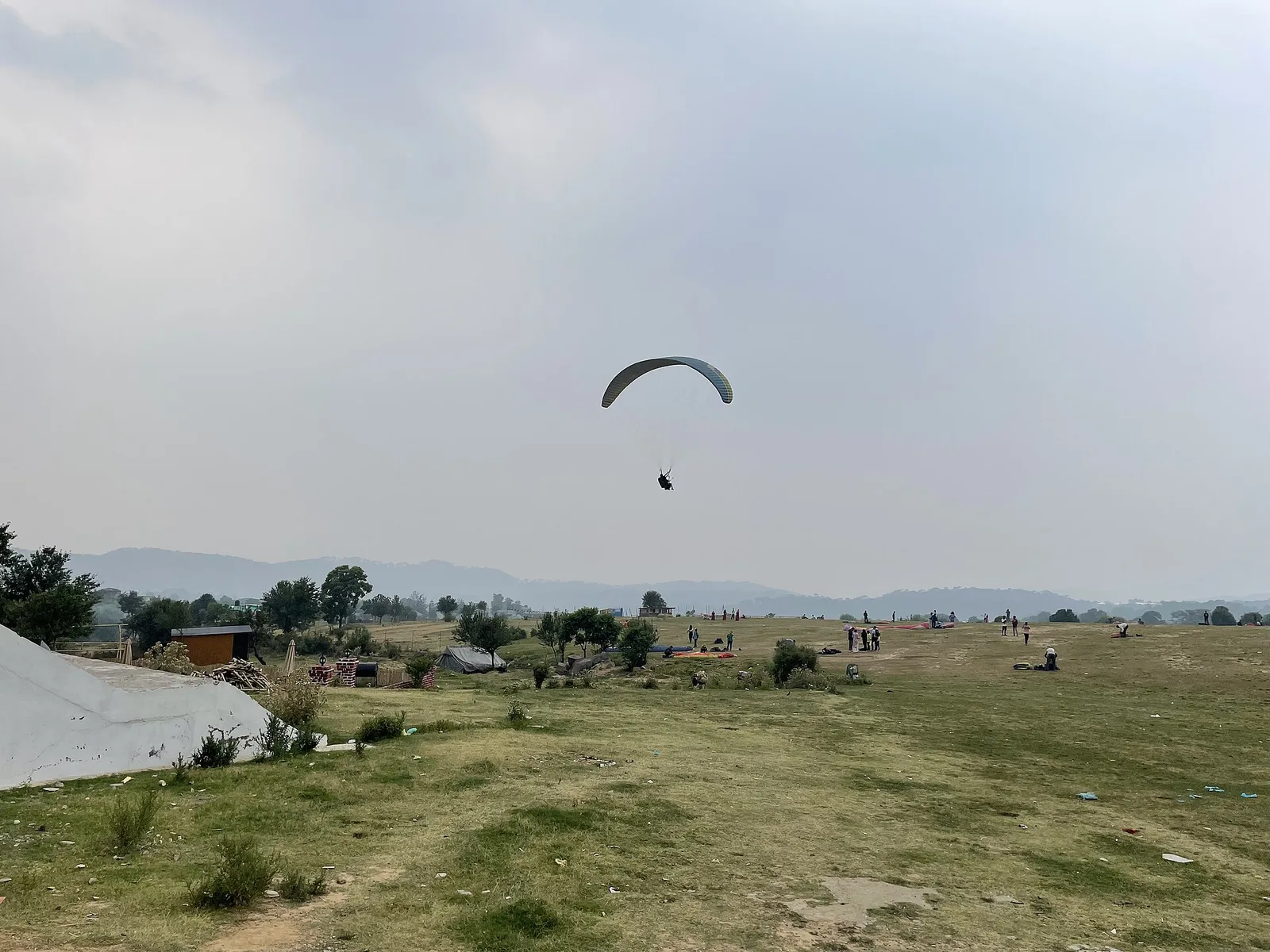A paraglider coming in to land over the open grassy landing ground at Bir with hills in the distance under a cloudy sky