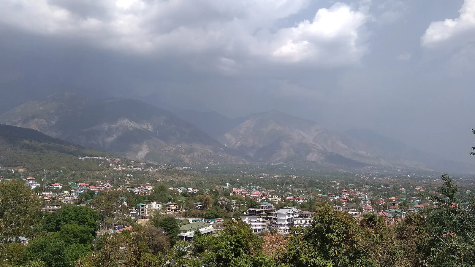 View over the town of Dharamshala with houses among green trees and the Dhauladhar mountains under heavy cloud