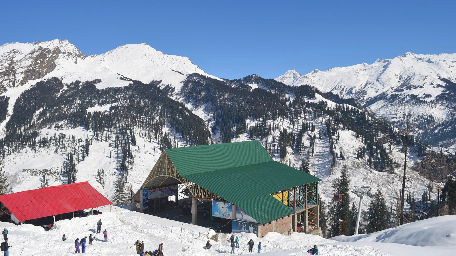 Snow-covered slopes and ski area buildings at Solang Valley near Manali under a clear blue sky, with visitors walking on the snow