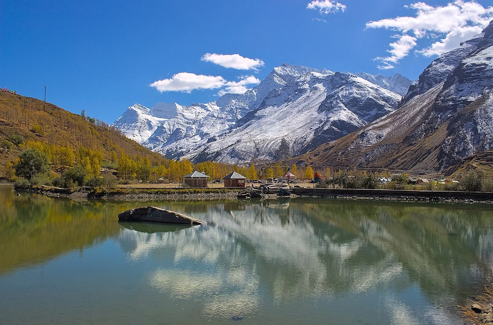 Sissu Lake in Lahaul with still water reflecting snow-capped peaks, yellow autumn trees on the shore and a few small buildings