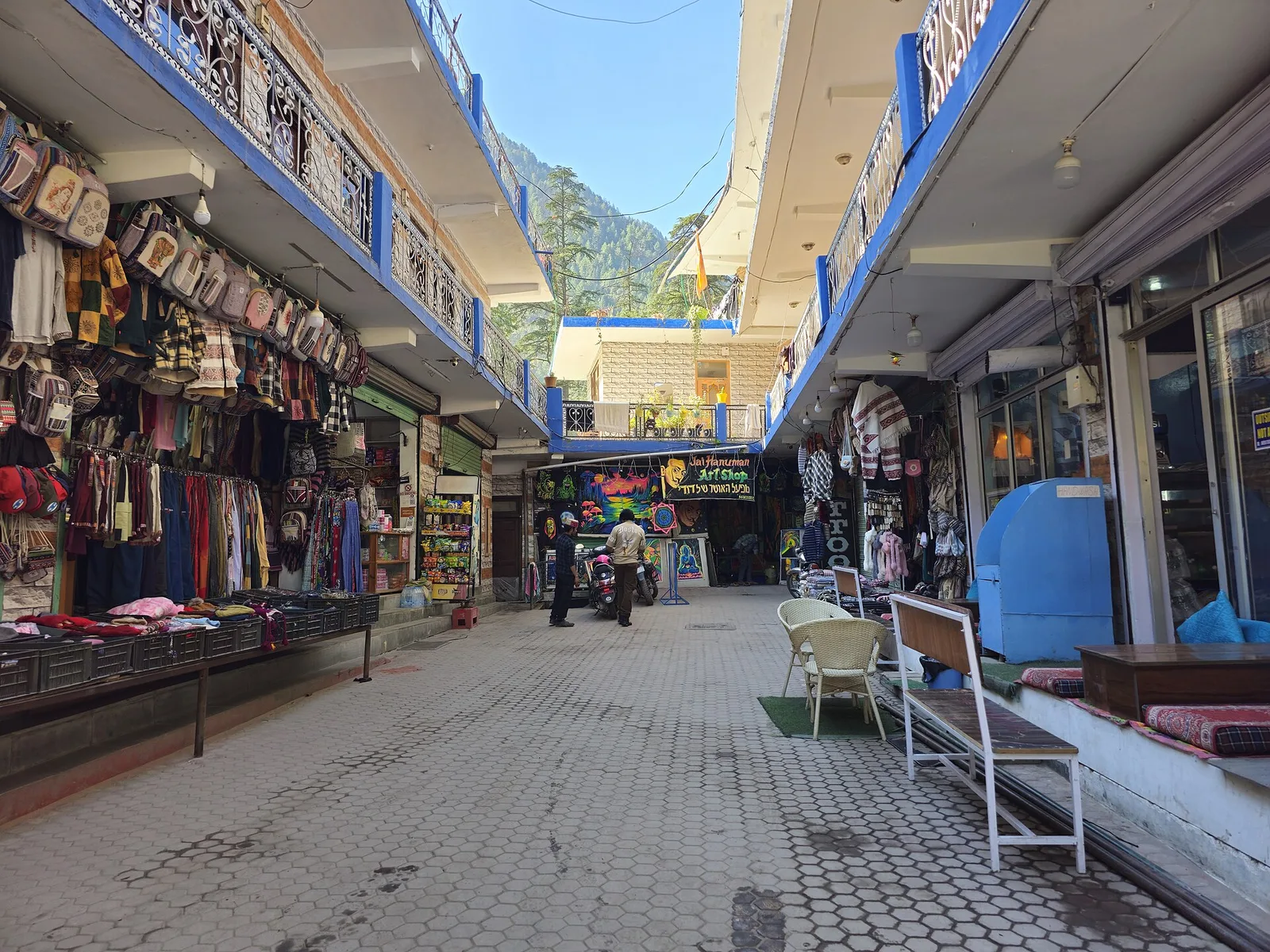 A paved market lane in Kasol lined with shops selling clothes and bags, with pine-covered hills and blue sky beyond