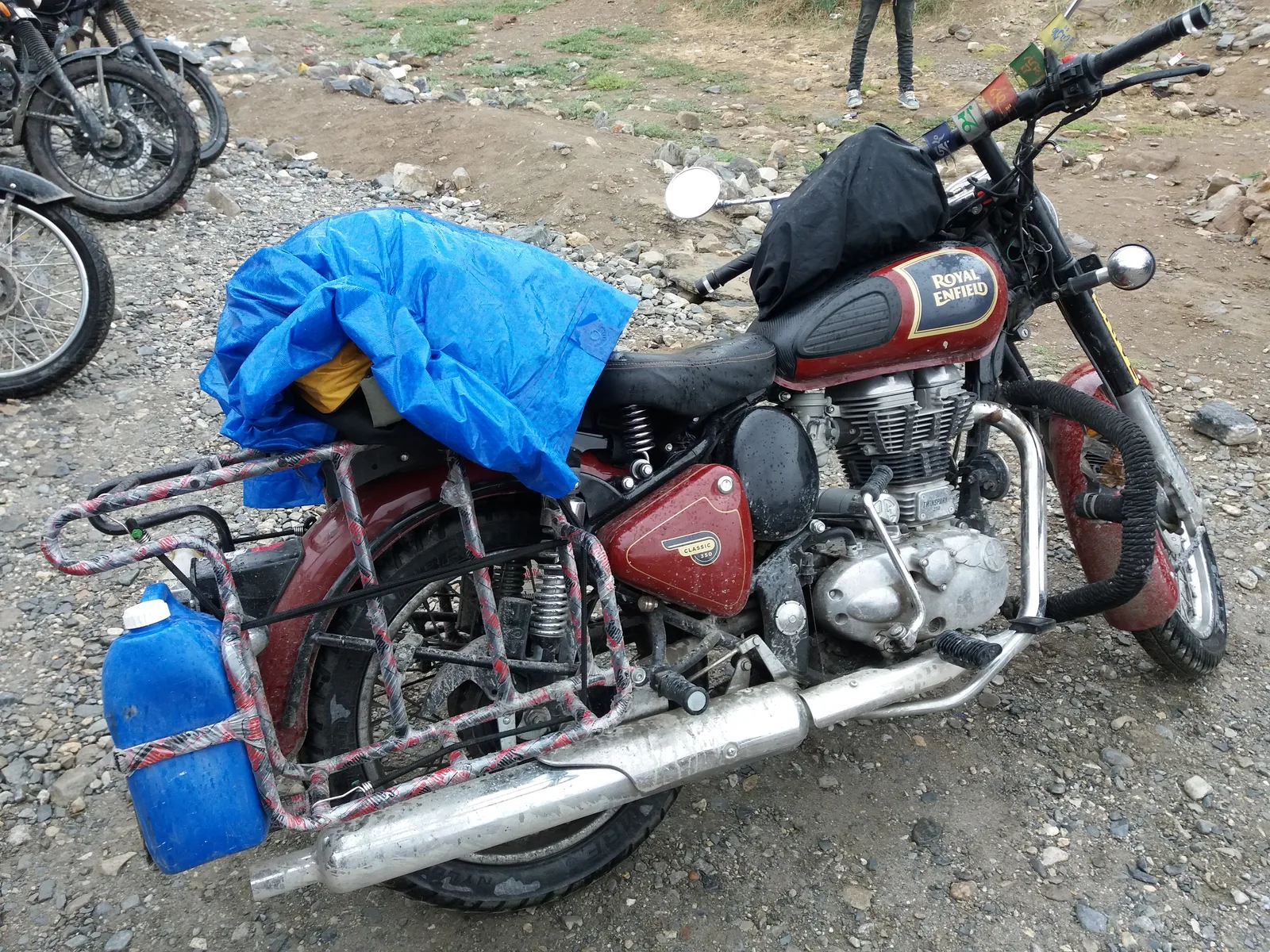 A red Royal Enfield Classic 350 loaded with a blue tarpaulin bag, a jerry can and pannier rails, parked on gravel beside the Leh to Manali highway
