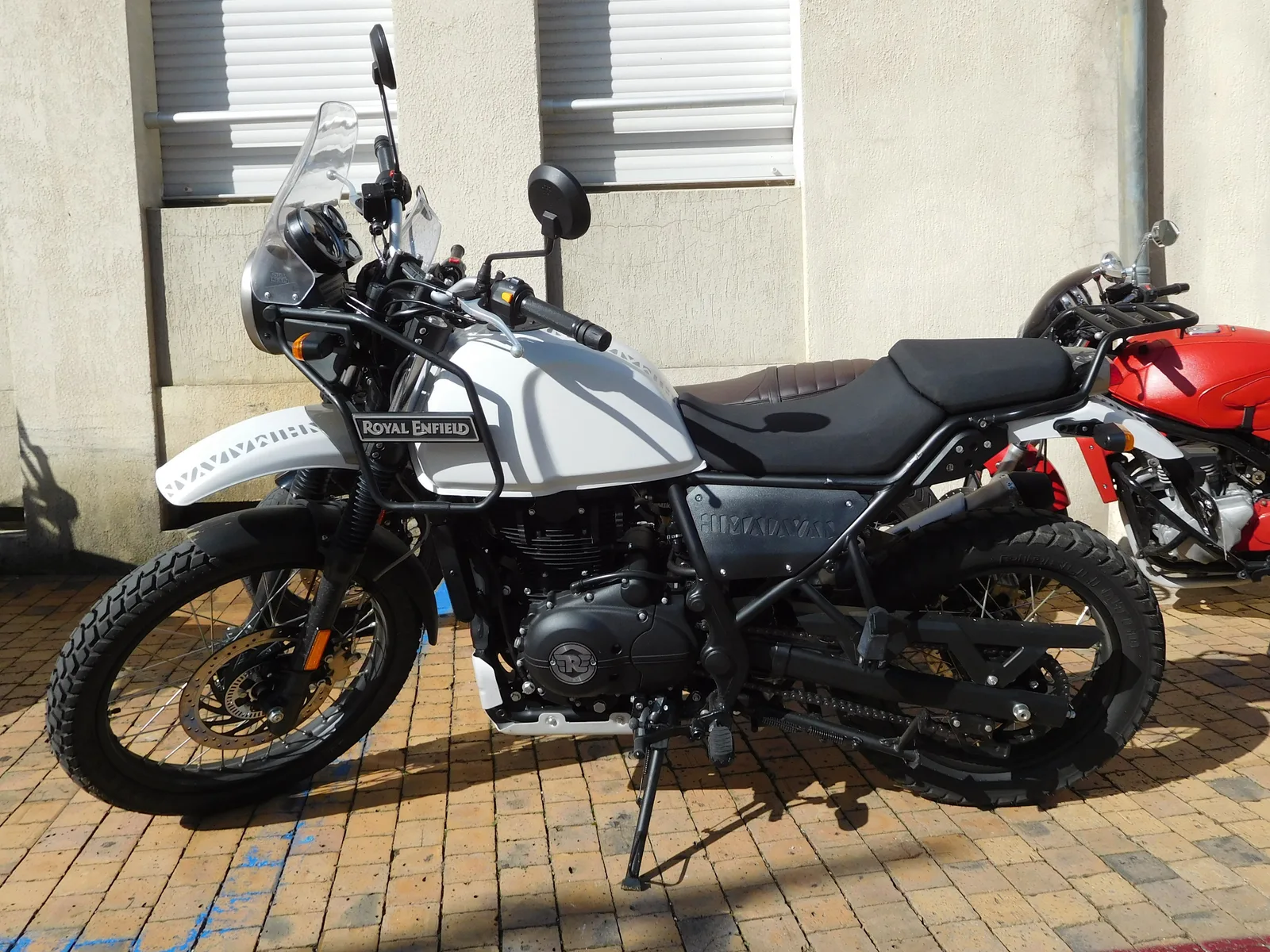 A white and black Royal Enfield Himalayan adventure motorcycle with a tall windscreen and spoked 21-inch front wheel, parked on a brick pavement