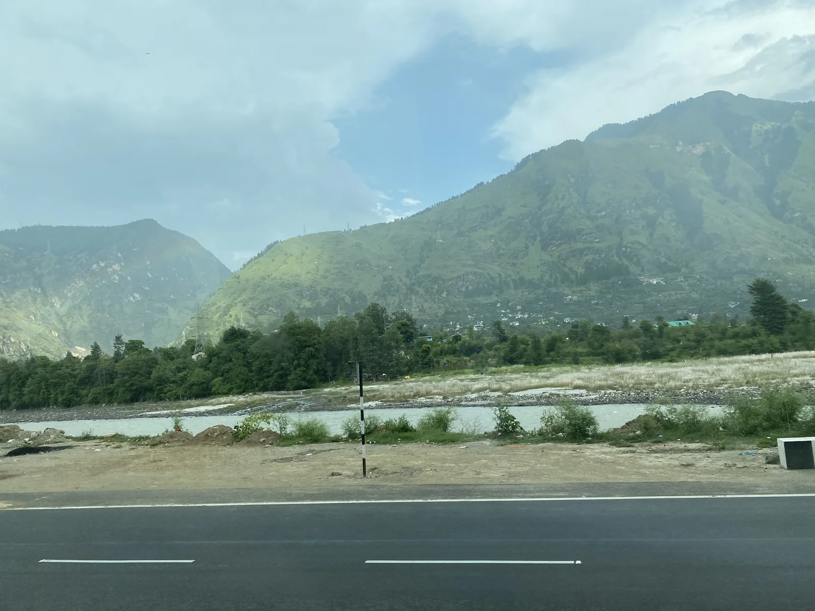 A wide, empty highway beside the shallow Beas river with green mountains under a cloudy sky, between Aut and Bhuntar