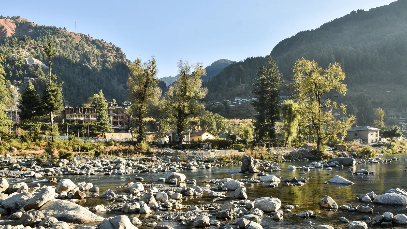 The rocky Uhl river at Barot in late October with autumn trees, village houses and pine-covered hills behind