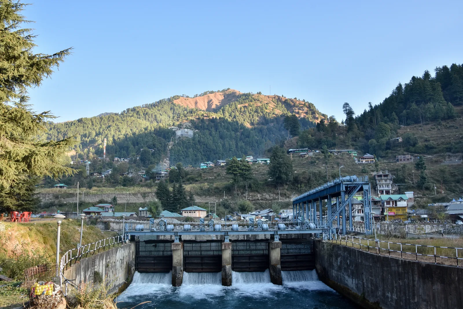 The concrete Uhl barrage at Barot releasing water, with the village and forested mountain behind under a clear sky