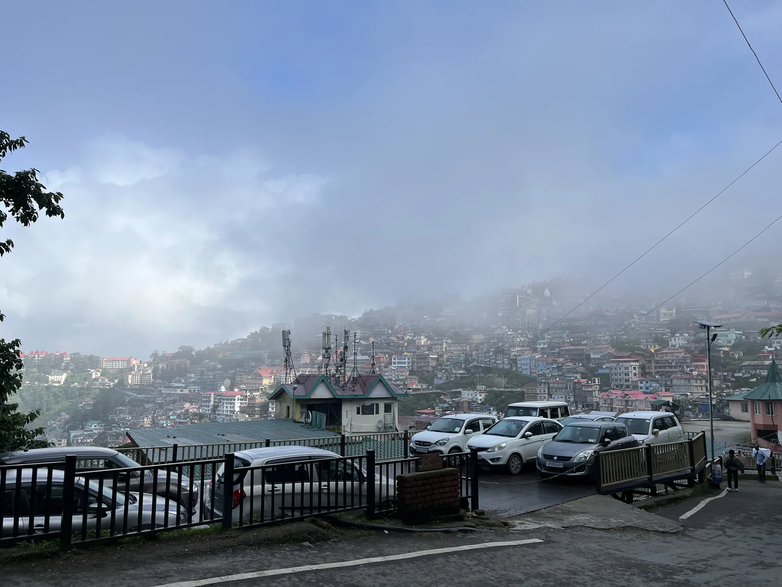 Cars parked behind a railing on a hillside road in Shimla, with the town spread across the slope below in mist