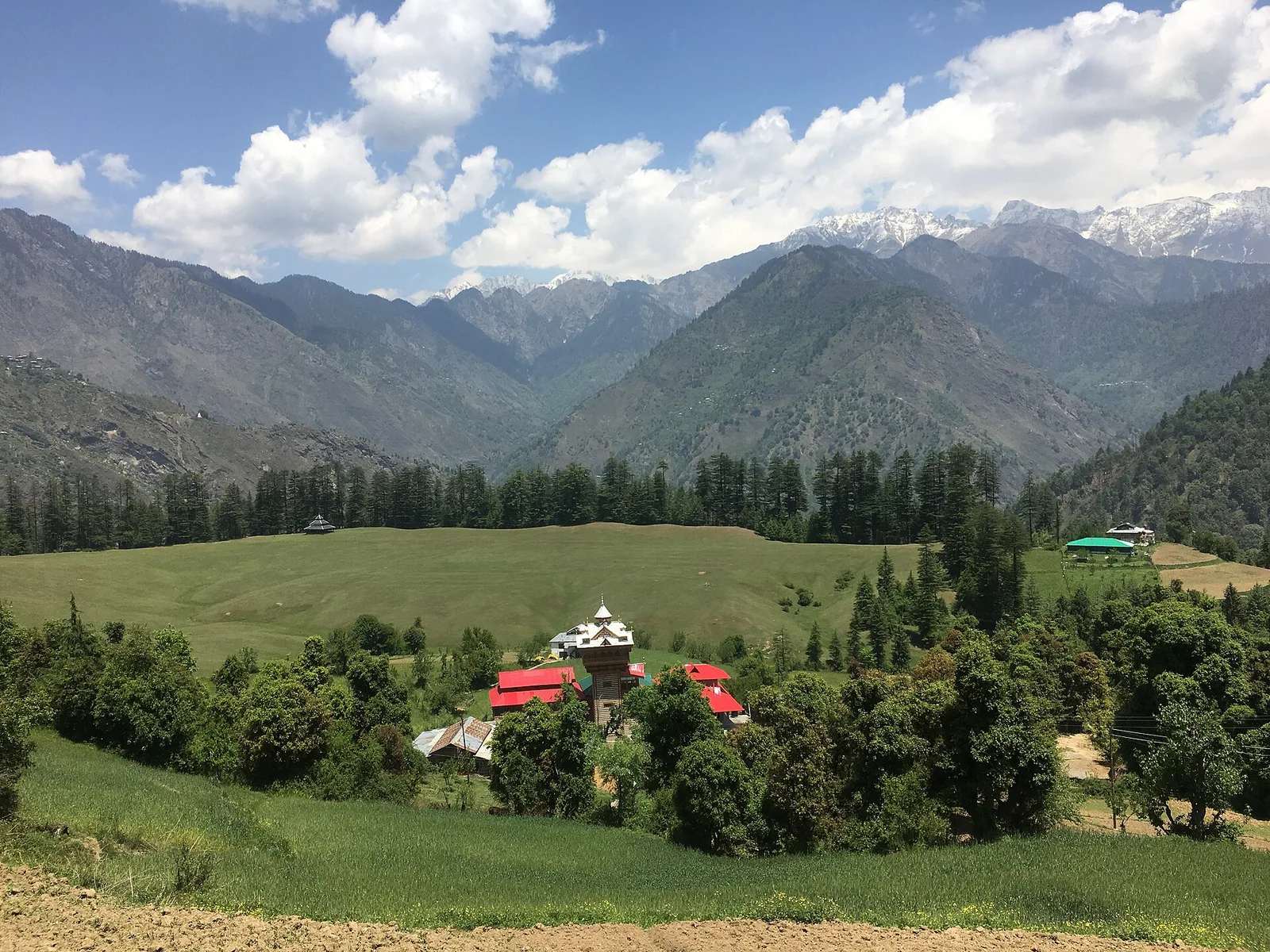 Shangarh village with red roofs and a pagoda-style temple among trees, the meadow in front and forested ridges behind