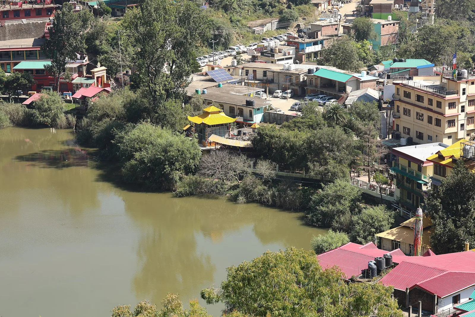 Rewalsar lake with trees along the shore, the town and parked cars on the slope above it